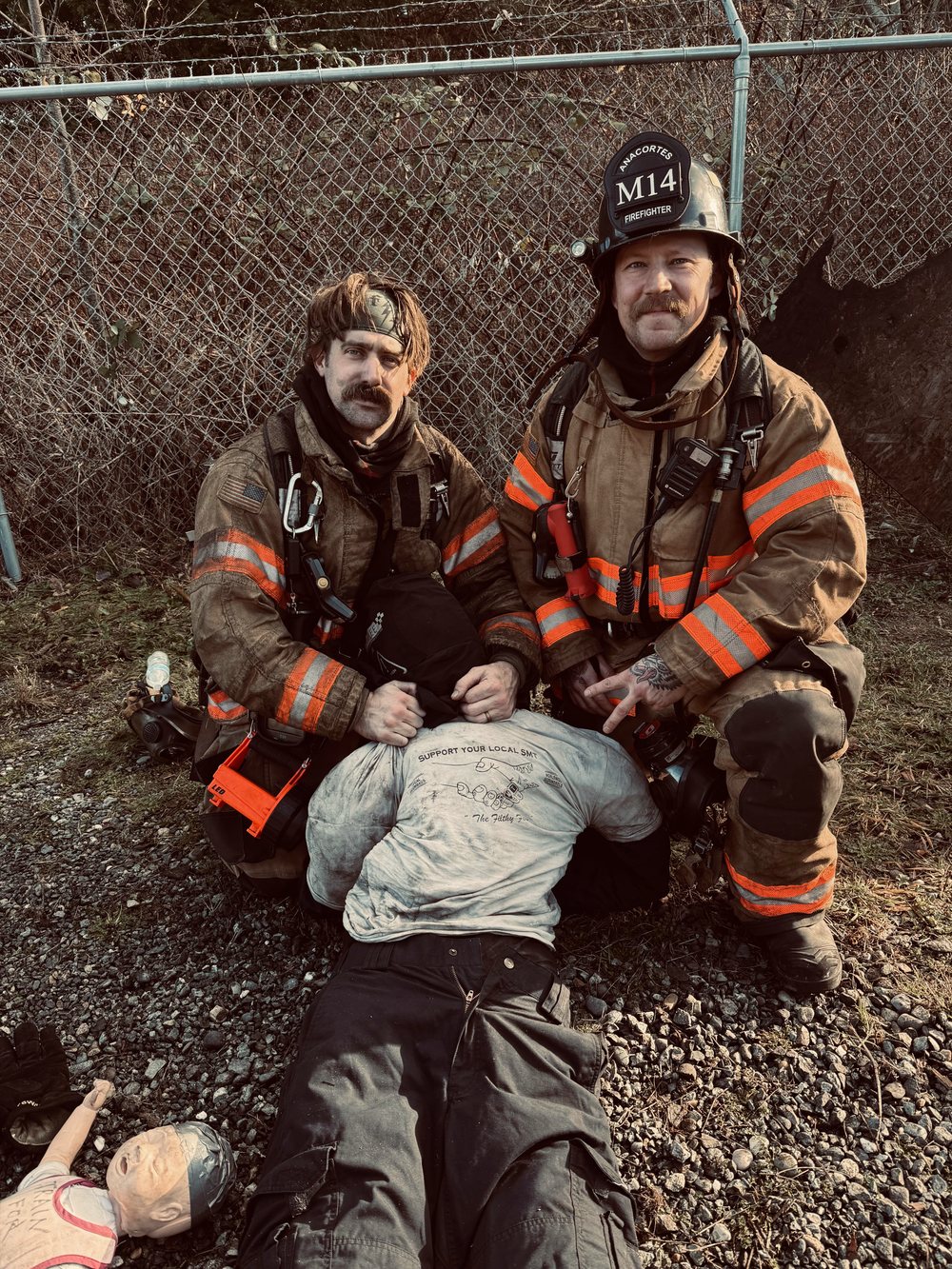 Two firefighters in soot-stained turnout gear kneeling behind a rescue training manikin after a training evolution. Both are former U.S. Navy Search and Rescue Medical Technicians now working as firefighter/paramedics.